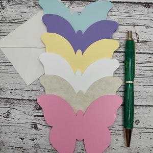 May include: A stack of six butterfly-shaped paper cards in light blue, purple, yellow, white, beige, and pink, accompanied by a white envelope and a green marbled ballpoint pen with gold-tone accents, arranged vertically on a wood-grain surface.