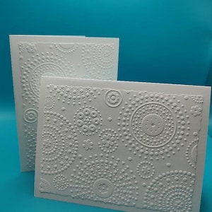 May include: Two white greeting cards with embossed dot and floral patterns. The cards have a textured surface and a deckled edge. The background is a solid teal color.