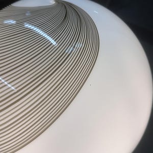 May include: A white, spherical glass lamp with a brown, swirling, striped design. The stripes curve across the surface, creating a dynamic visual effect. The lamp is illuminated, suggesting it is turned on.