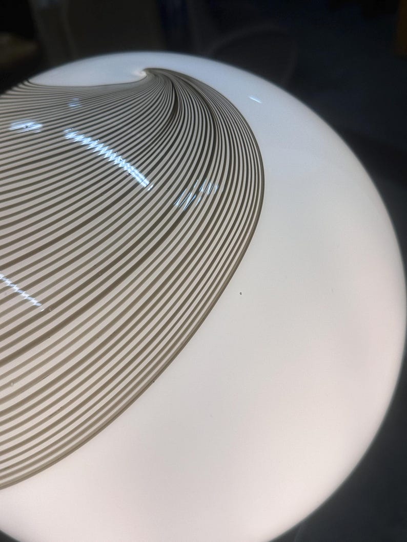 May include: A white, spherical glass lamp with a brown, swirling, striped design. The stripes curve across the surface, creating a dynamic visual effect. The lamp is illuminated, suggesting it is turned on.