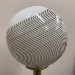 May include: A spherical lamp shade with a white top and gray striped bottom. The shade is mounted on a brass-colored base. The glass globe has a glossy finish.