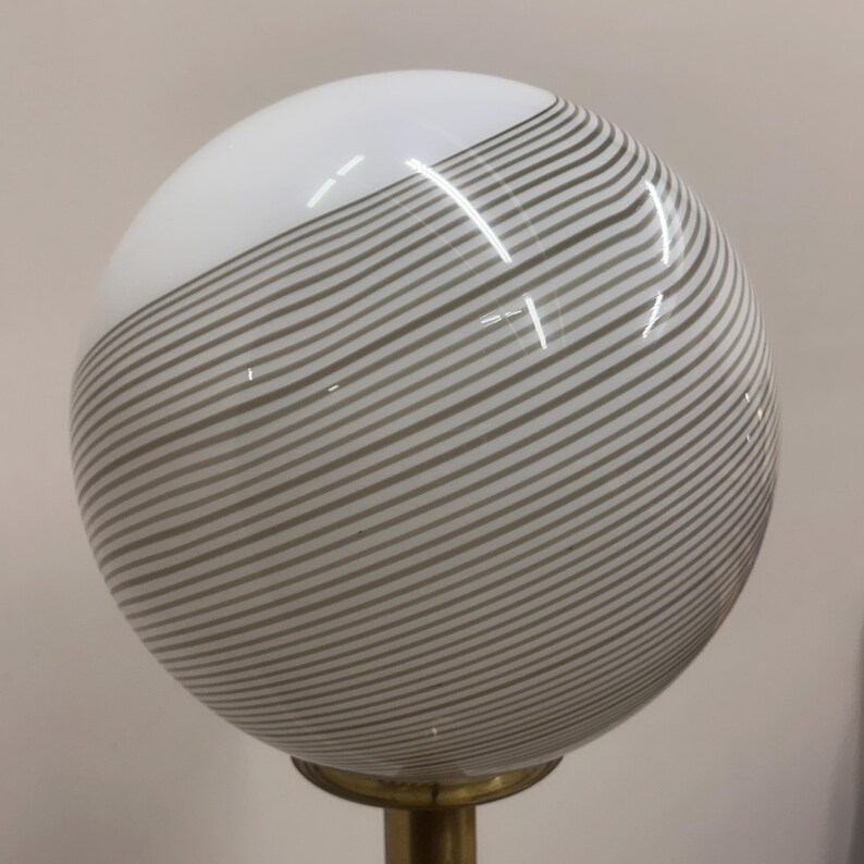 May include: A spherical lamp shade with a white top and gray striped bottom. The shade is mounted on a brass-colored base. The glass globe has a glossy finish.