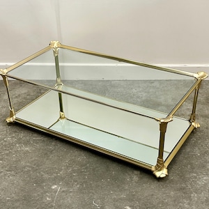 Könnte beinhalten: Ein goldfarbener Glas-Couchtisch mit einer verspiegelten unteren Ablage. Der Tisch hat eine rechteckige Form und verzierte Details an den Beinen.