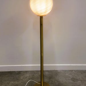 May include: A vintage floor lamp with a spherical, swirled glass shade. The shade sits atop a brass-colored pole and a circular base. The lamp is plugged into a wall outlet. The lamp is a decorative lighting fixture.