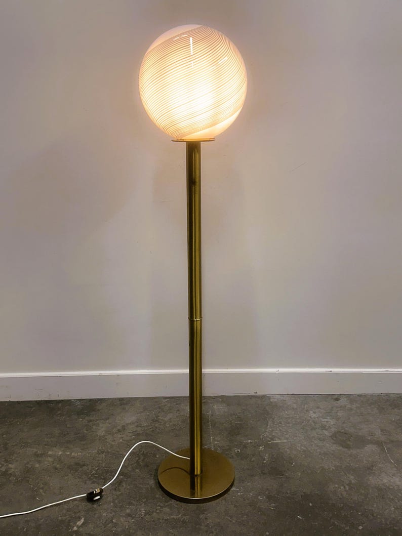 May include: A vintage floor lamp with a spherical, swirled glass shade. The shade sits atop a brass-colored pole and a circular base. The lamp is plugged into a wall outlet. The lamp is a decorative lighting fixture.