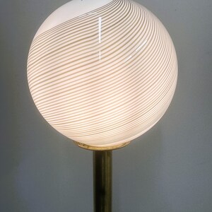 May include: A globe-shaped lamp with a white and beige striped pattern. The lamp sits atop a brass-colored pole. The light is on, illuminating the striped design. The lamp is a decorative lighting fixture.