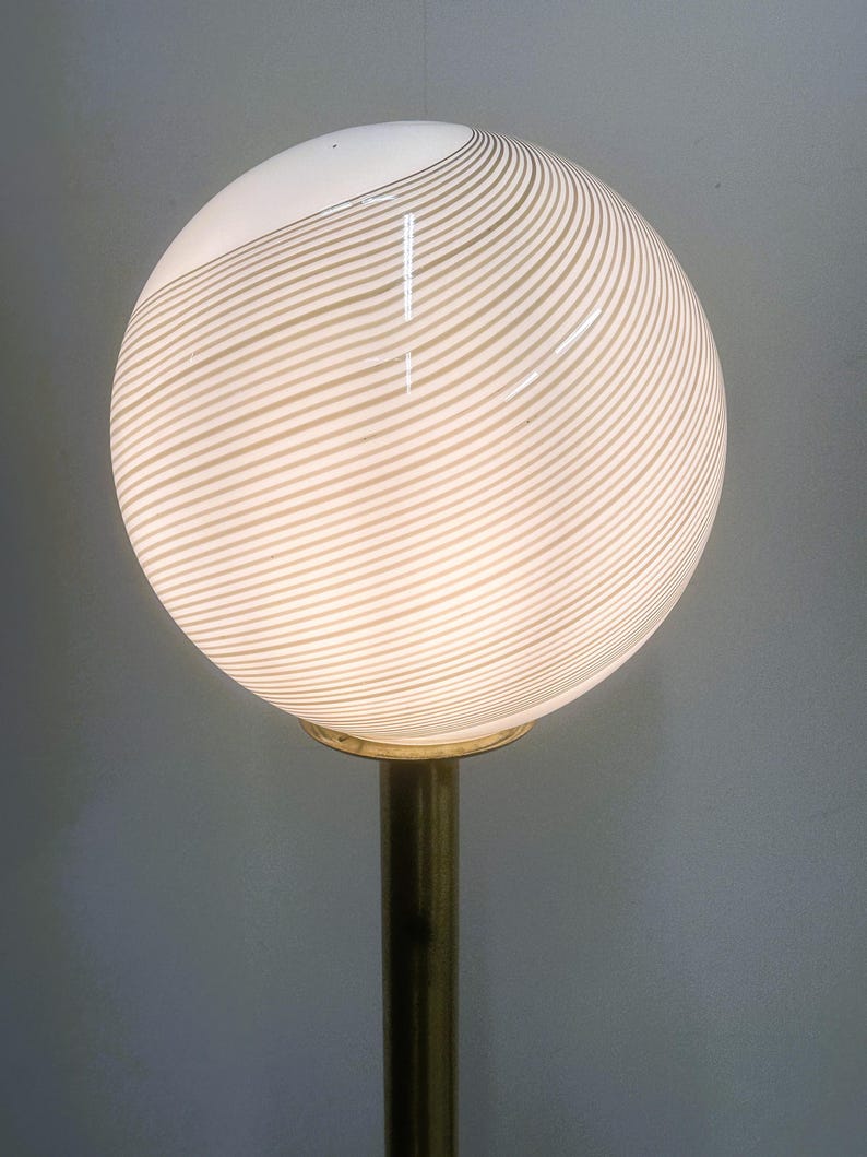 May include: A globe-shaped lamp with a white and beige striped pattern. The lamp sits atop a brass-colored pole. The light is on, illuminating the striped design. The lamp is a decorative lighting fixture.