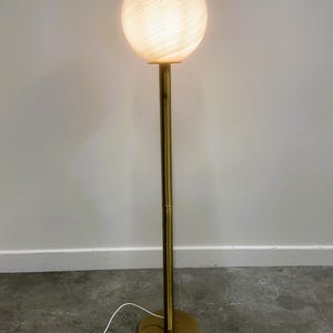 May include: A modern floor lamp with a spherical, swirled glass shade. The shade is illuminated, emitting a warm glow. The lamp has a slender, brass-colored metal pole and a circular base. The lamp is plugged into a wall outlet.