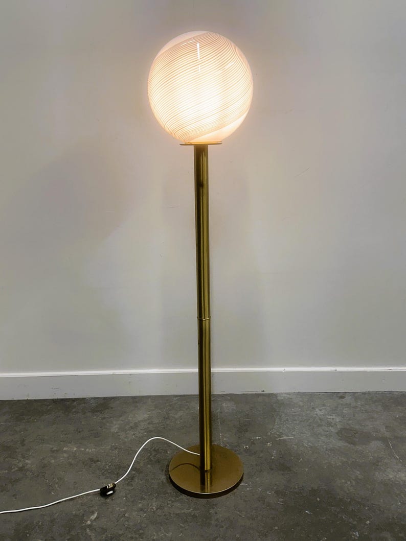 May include: A modern floor lamp with a spherical, swirled glass shade. The shade is illuminated, emitting a warm glow. The lamp has a slender, brass-colored metal pole and a circular base. The lamp is plugged into a wall outlet.