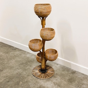 Può includere: Un portapiante alto a più livelli in rattan intrecciato. Il supporto presenta quattro fioriere rotonde a forma di ciotola e una base circolare. Il colore marrone naturale e la trama intrecciata gli conferiscono un'estetica vintage e bohémien.