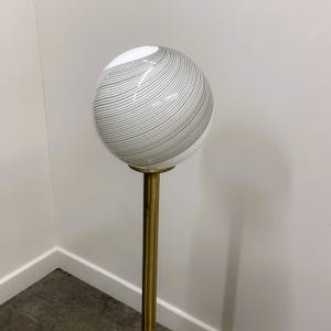 May include: A vintage floor lamp featuring a spherical glass shade with gray swirl patterns. The shade sits atop a brass-colored metal pole. The lamp is a decorative lighting fixture.