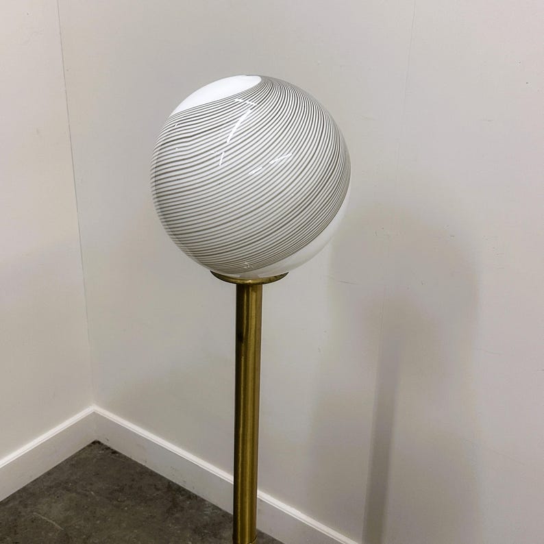 May include: A vintage floor lamp featuring a spherical glass shade with gray swirl patterns. The shade sits atop a brass-colored metal pole. The lamp is a decorative lighting fixture.