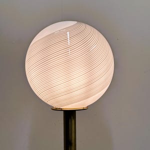 May include: A modern lamp featuring a large, spherical white glass shade with a striped pattern. The shade sits atop a brass-colored metal base. The lamp is designed to provide ambient lighting.