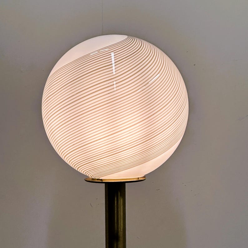 May include: A modern lamp featuring a large, spherical white glass shade with a striped pattern. The shade sits atop a brass-colored metal base. The lamp is designed to provide ambient lighting.