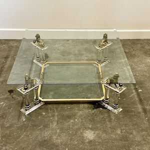 May include: A square glass coffee table with a gold-colored metal frame and clear acrylic legs. The legs are topped with decorative lion figures. The table has a lower shelf and is suitable for a living room.