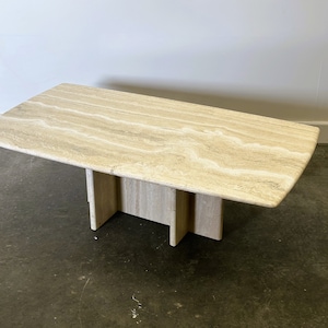 Pode incluir: Uma mesa de centro retangular de travertino de cor bege claro com um padrão ondulado único. A mesa tem um design simples e moderno com quatro pernas que fazem parte da superfície da mesa.