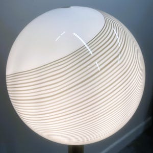 May include: A spherical lamp with a white top and a lower section with horizontal, wavy, beige stripes. The lamp is mounted on a dark, cylindrical base. The lamp's design is modern and minimalist.
