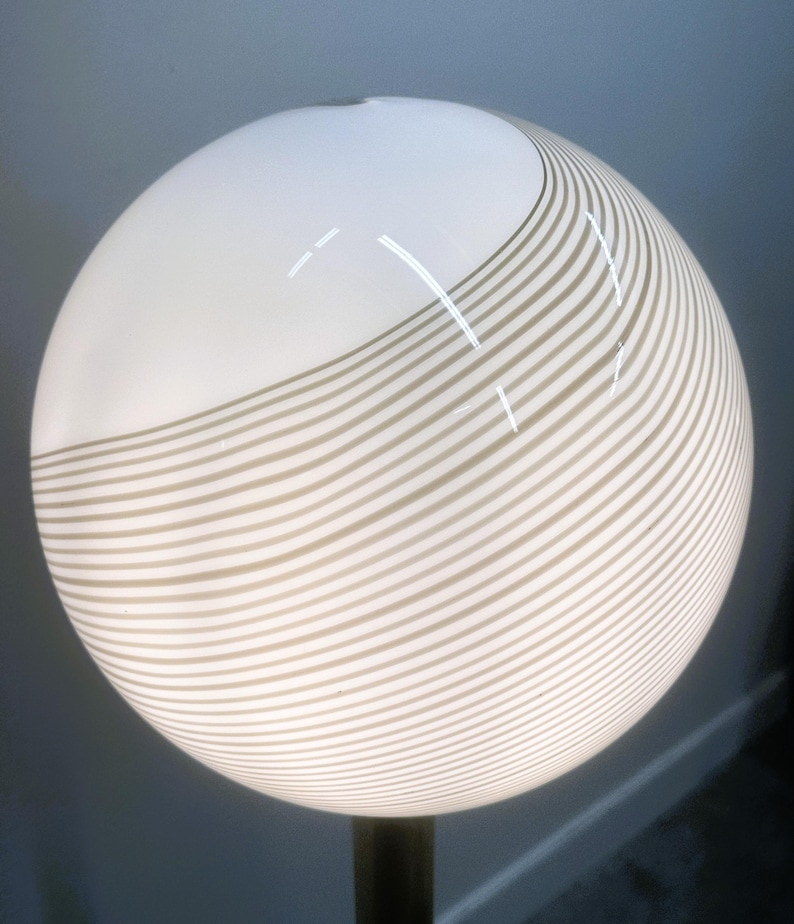 May include: A spherical lamp with a white top and a lower section with horizontal, wavy, beige stripes. The lamp is mounted on a dark, cylindrical base. The lamp's design is modern and minimalist.
