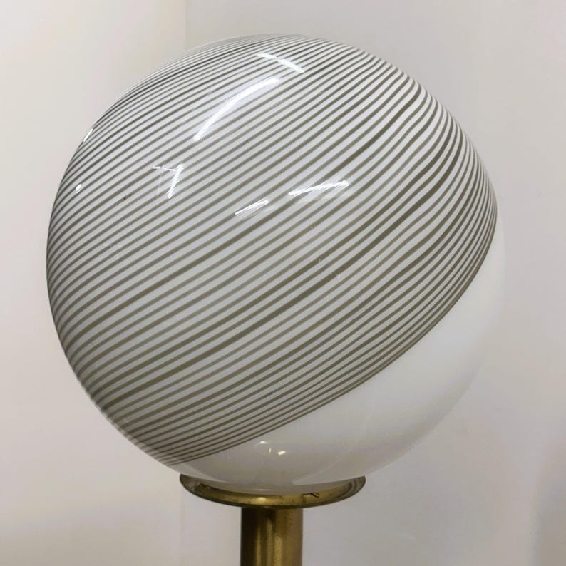 May include: A spherical lamp with a white and gray striped glass shade. The shade is mounted on a brass-colored metal base. The lamp has a modern design and is suitable for home decor.
