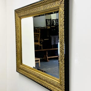 May include: A large, rectangular mirror with a black frame and a gold-colored ornate border. The mirror is reflecting a room with furniture.