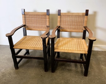May include: Two matching armchairs with woven cane seats and backs. The chairs have dark wooden frames and curved armrests. The woven cane is a light tan color, contrasting with the dark wood. The chairs are designed for seating.