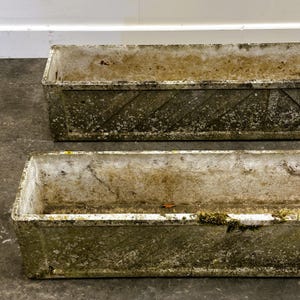 Peut inclure: Deux jardinières rectangulaires en béton vieilli. Les jardinières présentent une surface rugueuse et texturée avec des signes de vieillissement, notamment de la mousse et une décoloration. Le design comprend un motif en diagonale sur les côtés.