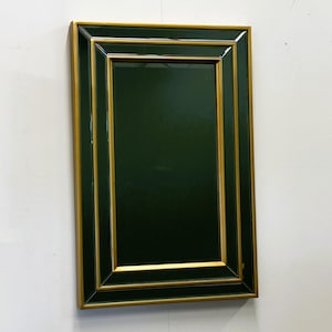 Può includere: Specchio da parete rettangolare con centro verde scuro e cornice dorata. La cornice ha un design a strati, che aggiunge profondità all'aspetto dello specchio. Lo specchio è montato su una parete bianca.