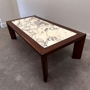 Può includere: Tavolino rettangolare con piano in marmo e struttura in legno scuro. Il marmo presenta un motivo bianco e grigio. Il tavolo ha un profilo basso con quattro gambe. Dimensioni: circa 120 cm di lunghezza, 60 cm di larghezza e 40 cm di altezza.