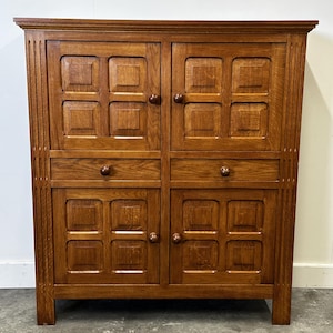 Könnte beinhalten: Ein mittelbrauner Holzschrank mit vier Türen und zwei Schubladen. Die Türen haben eingelassene quadratische Paneele und runde Holzknöpfe. Der Schrank hat eine rechteckige Form und ist aus massivem Holz gefertigt, das eine natürliche Holzmaserung aufweist.