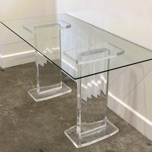 May include: A clear acrylic console table with a rectangular glass top. The table has two clear acrylic legs with a zig-zag design. The legs are attached to a rectangular base. The table is approximately 122 cm long.