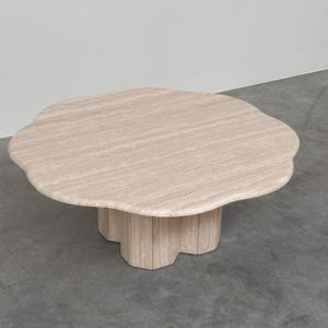 Può includere: Un tavolino basso realizzato in pietra travertino beige. Il piano del tavolo presenta una forma organica arrotondata simile a un fiore con bordi morbidi e smerlati. È sostenuto da una base centrale scanalata che riprende il design curvo.