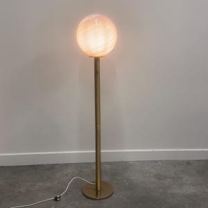 May include: A modern floor lamp with a round, glowing globe shade. The shade has a textured, swirled pattern and emits a warm light. The lamp has a slender, gold-colored metal pole and a circular base. The lamp is plugged into an electrical outlet.