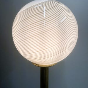 May include: A spherical lamp with a white and beige striped pattern. The globe-shaped lampshade sits atop a brass-colored metal base. The lamp is lit, emitting a soft glow.