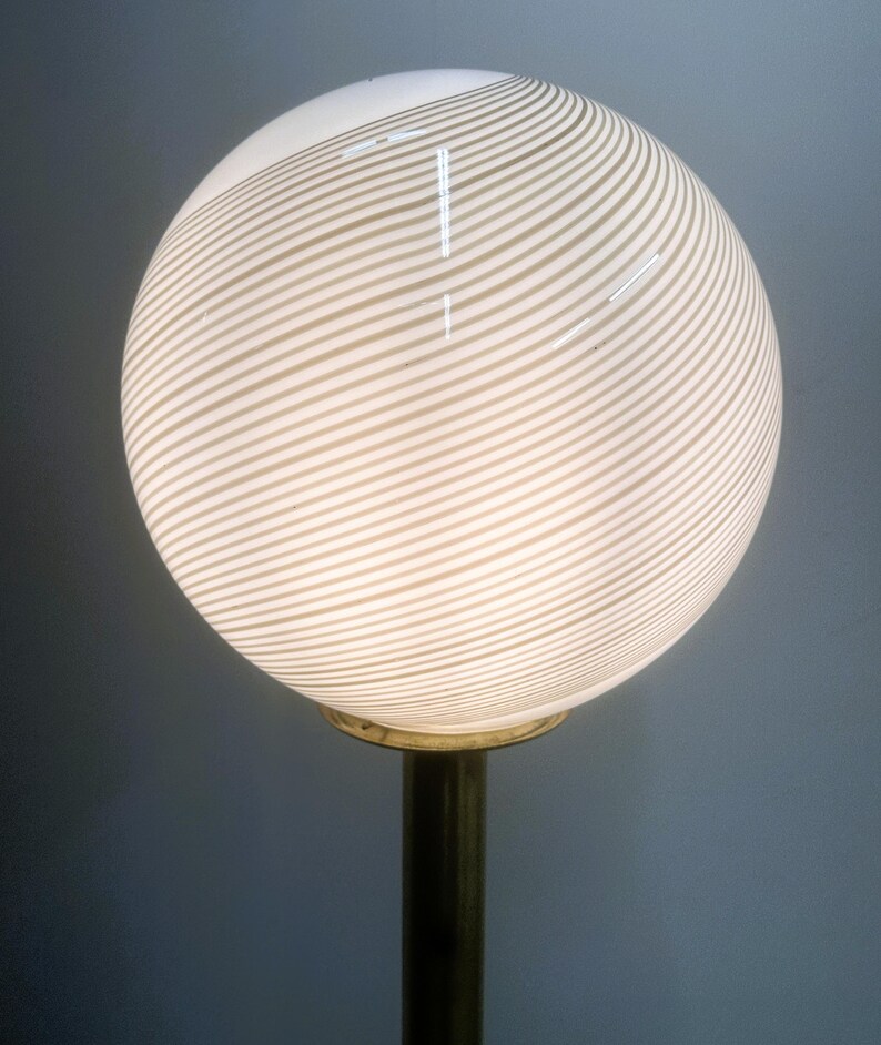 May include: A spherical lamp with a white and beige striped pattern. The globe-shaped lampshade sits atop a brass-colored metal base. The lamp is lit, emitting a soft glow.