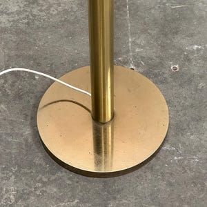 May include: A gold-colored floor lamp base with a round, flat base and a cylindrical pole. The base is circular, and the pole is centered. A white electrical cord extends from the base.