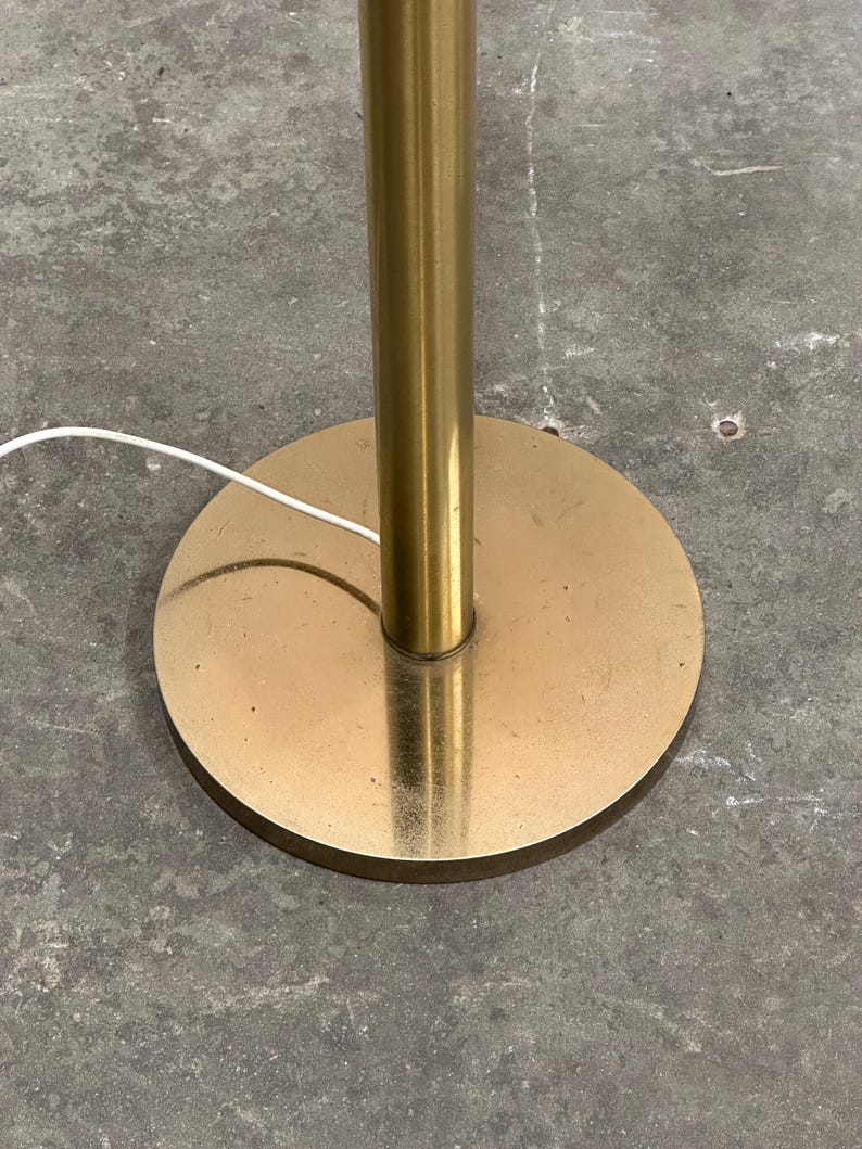 May include: A gold-colored floor lamp base with a round, flat base and a cylindrical pole. The base is circular, and the pole is centered. A white electrical cord extends from the base.
