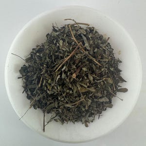 May include: A white bowl filled with dried green leaves and stems. The leaves are a dark green color and appear to be dried herbs.