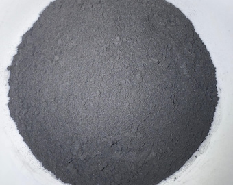 May include: A close-up image of a pile of fine, gray powder. The powder is densely packed, with a slightly uneven surface. The color is a uniform, muted gray, suggesting a mineral or industrial substance. The powder is contained within a white, rounded container.