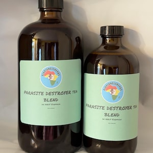 May include: Two brown glass bottles with black caps. Each bottle has a light green label with the text "PARASITE DESTROYER TEA BLEND DR SEBIS' FORMULA" and a circular logo. The bottles are filled with a dark liquid.