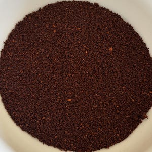 May include: A close-up shot of finely ground, dark brown coffee beans filling a white bowl. The coffee grounds have a rich, deep color and a slightly coarse texture, suggesting a fresh grind.