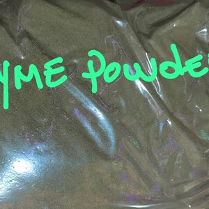 May include: A clear plastic bag filled with thyme powder. The words "Thyme Powder" are written in bright green, stylized letters across the top of the bag. The powder appears to be a dark green color.