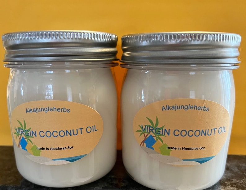 Op de afbeelding: Twee glazen potten met zilveren deksels gevuld met witte virgin kokosolie. De potten hebben gele etiketten met de tekst "Alkajungleherbs Virgin Coconut Oil made in Honduras 8oz."