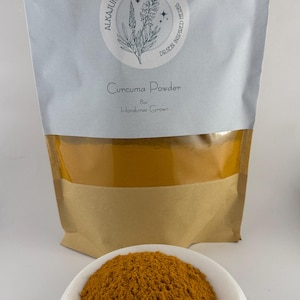 Może przedstawiać: Brązowa papierowa torba z białą etykietą, na której widnieje napis "AlkaJungleHerbs Dr. Sebi Inspired Herbs" oraz "Curcuma Powder 8oz Honduran Grown". Torba jest wypełniona jasno pomarańczowym proszkiem kurkumy. Na pierwszym planie znajduje się mała biała miseczka wypełniona proszkiem kurkumy.