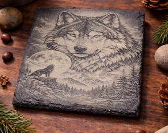 Puede incluir: Un posavasos cuadrado de pizarra gris oscuro con un grabado detallado de la cabeza de un lobo y un lobo aullando en un paisaje montañoso. El posavasos está rodeado de elementos naturales.