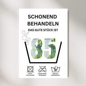 Puede incluir: Un póster blanco con texto en alemán y símbolos de lavado. El texto dice "SCHONEND BEHANDELN" y "DAS GUTE STÜCK IST" con el número "85" en el centro, que parece un billete de euro. Instrucciones de lavado en la parte inferior.