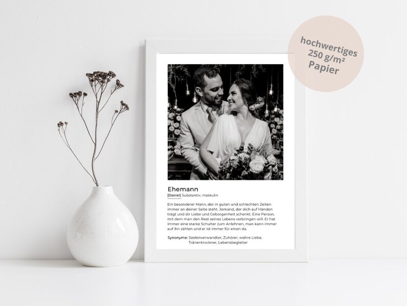 May include: A framed black and white photograph of a couple embracing, with text below describing the term "husband." The print is displayed on a white surface, with a white vase holding dried flowers to the left.