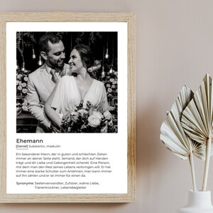 May include: A framed black and white photograph of a couple embracing, with the word "Ehemann" and a definition in German. The frame is light brown, and the background is a neutral color. The image also includes decorative dried palm leaves.