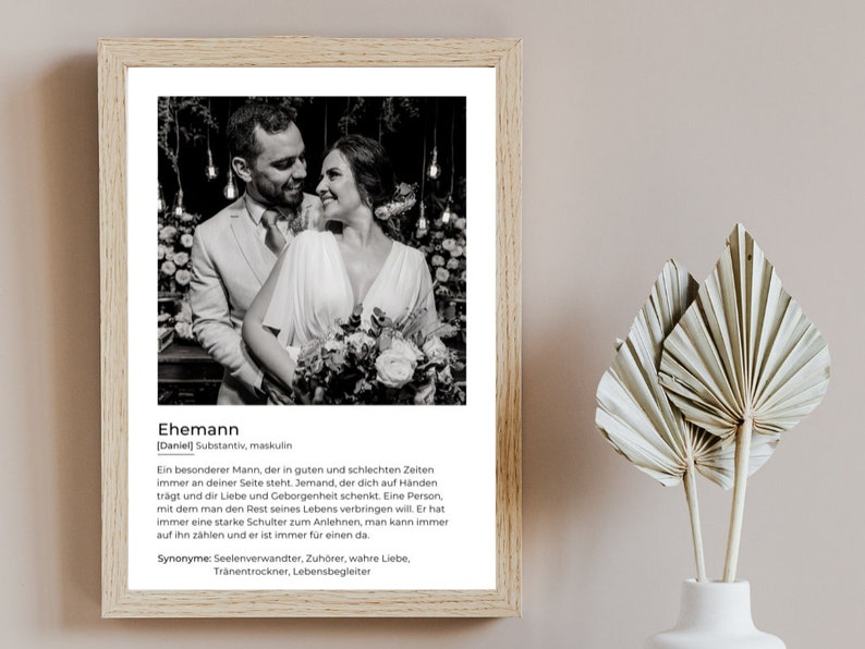 May include: A framed black and white photograph of a couple embracing, with the word "Ehemann" and a definition in German. The frame is light brown, and the background is a neutral color. The image also includes decorative dried palm leaves.
