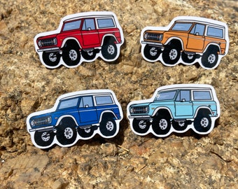 May include: Four different colored Ford Bronco stickers. The stickers are red, orange, blue, and light blue. Each sticker has a white roof and white trim around the windows.