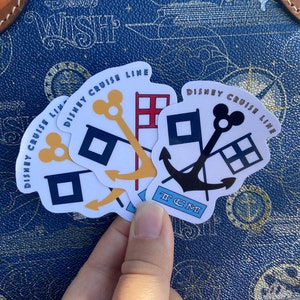 May include: Four white stickers with a blue anchor, a red and white checkered flag, a yellow key, and a black Mickey Mouse head. The stickers are arranged in a stack and say "Disney Cruise Line" in a gold and black font.